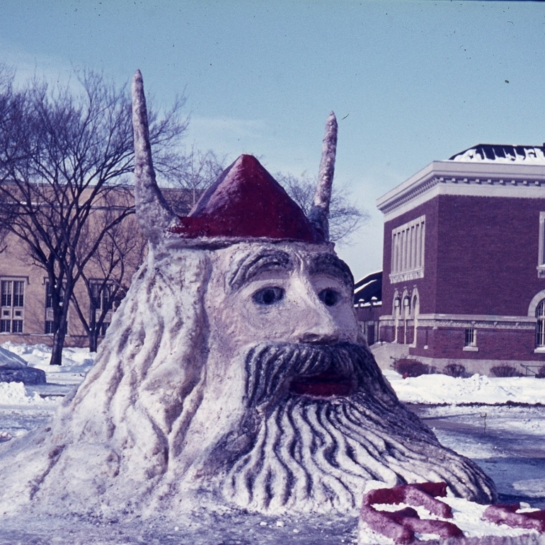 A snow sculpture of a Viking's head wearing a helmet with horns. The snow 'painted' with color for the red helmet, eyes, and dark beard. Archival picture showing two since razed campus buildings